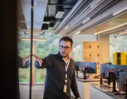 A man wearing glasses and a lanyard points to a reflective surface in a modern office with multiple computer monitors.