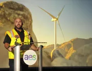 A man in a yellow safety vest speaks at a podium with the AES logo. Behind him, a large screen displays a wind turbine set against a mountainous landscape.