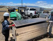 Two workers in hard hats and gloves handle a stack of solar panels on a pallet at a solar farm. A green tractor and a white truck are parked nearby. Rows of solar panels stretch into the distance under a blue sky.