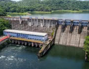 Aerial view of the Bayano Dam surrounded by lush green forests. The dam features a large concrete structure with multiple spillways and a building labeled 'BAYANO'. The reservoir is calm, and water flows out from the dam into the river below.