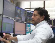 A man in a white shirt sits at a desk surrounded by multiple computer monitors displaying various data and graphs.