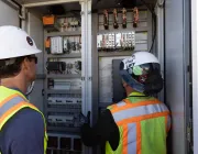 Two construction workers wearing safety vests and helmets are inspecting an open electrical panel with various equipment and wiring inside.