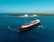 A large LNG tanker is escorted by two tugboats on a vast blue ocean. In the background, an island with green vegetation and white storage tanks is visible under a clear blue sky.