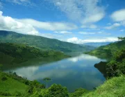 A scenic view of a lake surrounded by lush green hills under a partly cloudy sky. The water reflects the sky and clouds, creating a serene and picturesque landscape.
