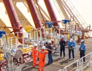 A group of workers in safety gear stand on a metal platform beside large industrial pipes and valves on a ship. The pipes are red and yellow, with blue and yellow valve controls. The workers are engaged in discussion, with some looking at a device. A ladder and railing are visible in the background.