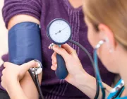 A healthcare professional measures a patient's blood pressure using a sphygmomanometer. The patient wears a purple sweater, and the professional holds the device's gauge and bulb while listening with a stethoscope.