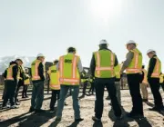 Construction workers wearing orange safety vests and helmets stand and converse on a gravel site with portable buildings in the background. Sunlight creates a lens flare effect.