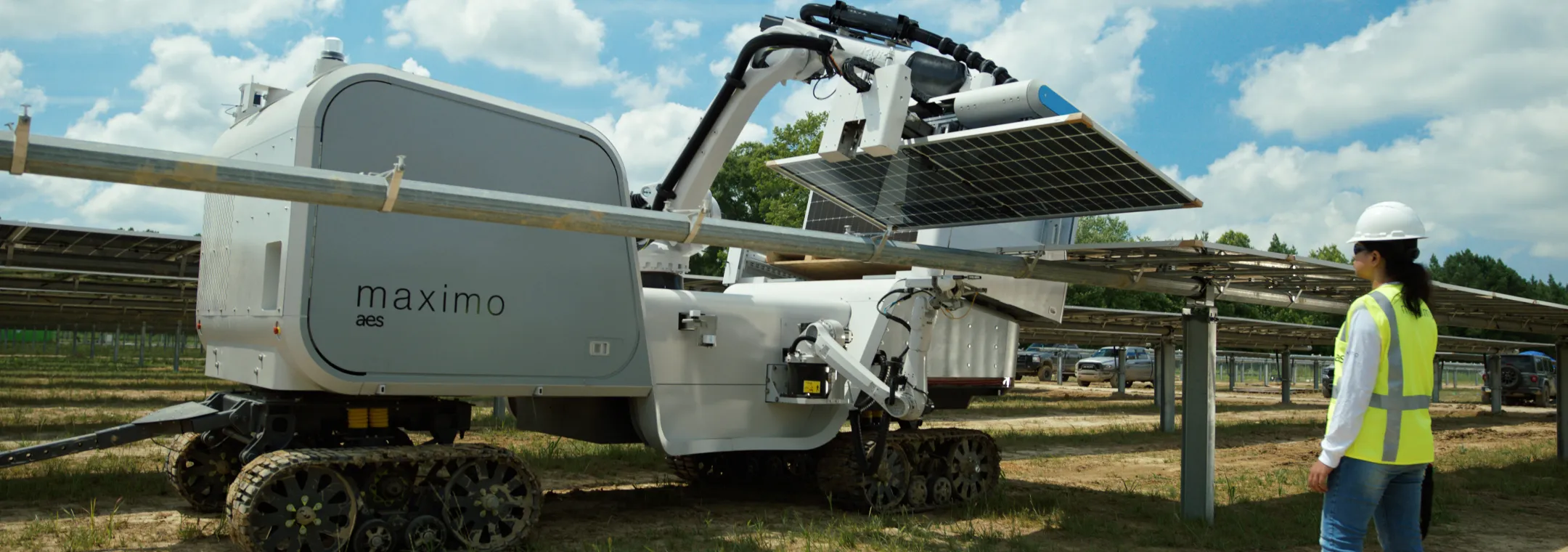 A robotic machine with tracks, labeled Maximo, is installing solar panels in a field. A person wearing a hard hat and high-visibility vest observes the process.