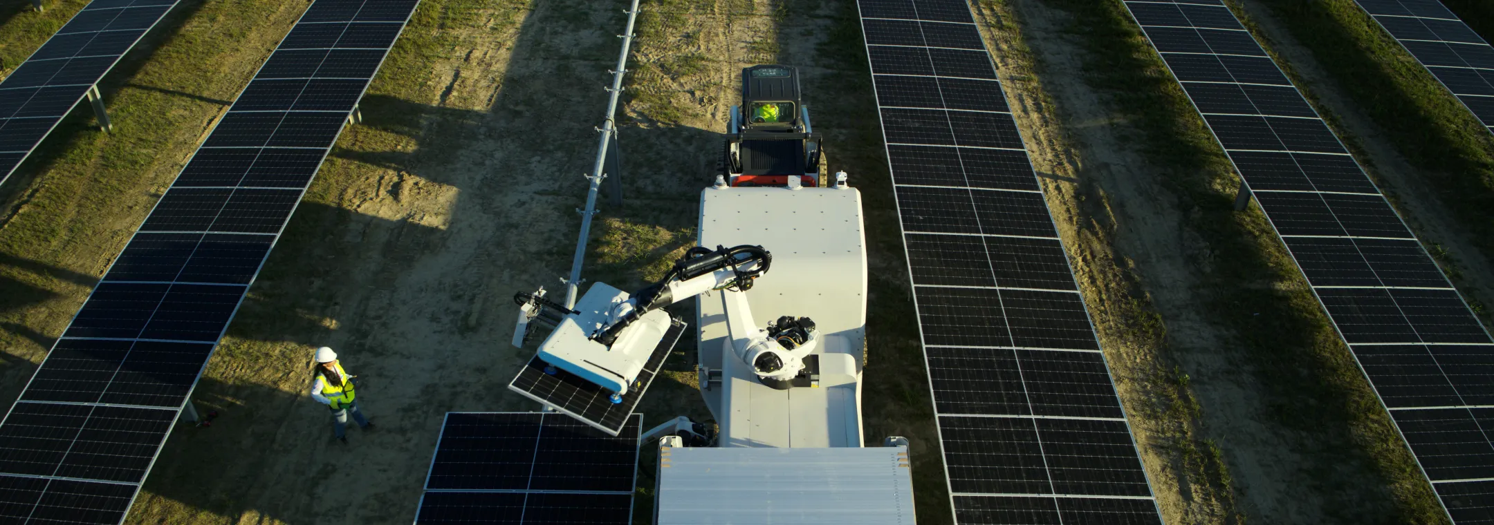 Aerial view of a robotic arm cleaning solar panels in a solar farm, with a worker in a safety vest and helmet observing nearby.