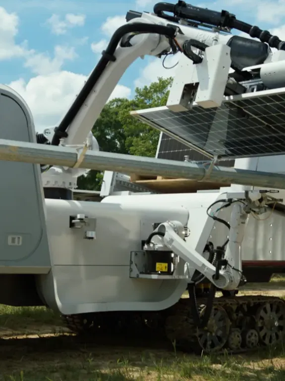 A large robotic machine with tracks is adjusting solar panels in a field. A person wearing a hard hat and reflective vest observes the process. The sky is partly cloudy.